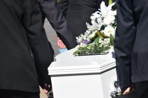 Personas de luto en trajes negros junto a un ataúd blanco adornado con flores blancas y moradas, mostrando un ambiente solemne y de respeto
